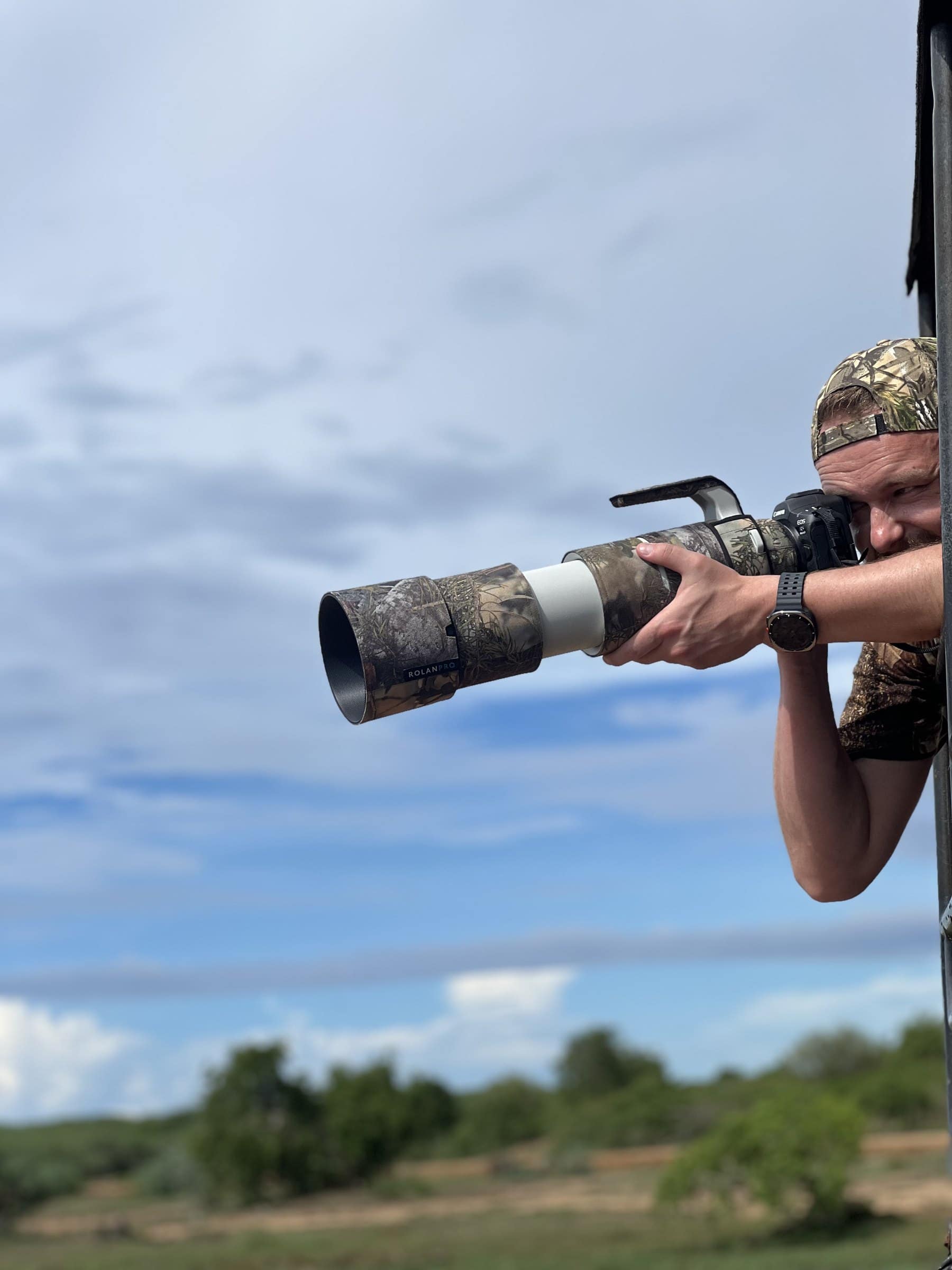 A wildlife photographer leaning out of a safari jeep with a camouflaged telephoto lens